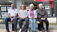 A group of adults, male and female, sat on a street bench smiling