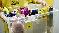A close up of a person wearing a yellow Boots t shirt carrying a clear plastic box with tolietries