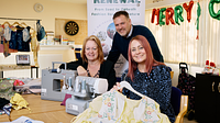 Two ladies sat beside a sewing machine and garments and a man looking over their shoulders