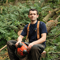 A man sat in the woods holding an orange safety helmet