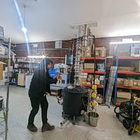 Elliot Rose standing next to a gin distilling machine in a red brick warehouse building surrounded by shelves of glass bottles and equiptment.