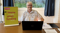 A man sat behind a desk wearing glasses smiling, the desk has papers and leaflets